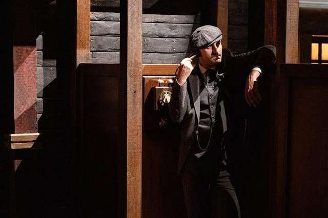 Man in flat cap and dark suit posing in vintage telephone booth with period street setting.