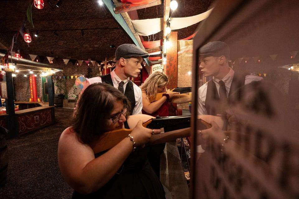 Indoor venue with brick walls, string lights, and themed decor. Guests in period costume interact at a bar counter.
