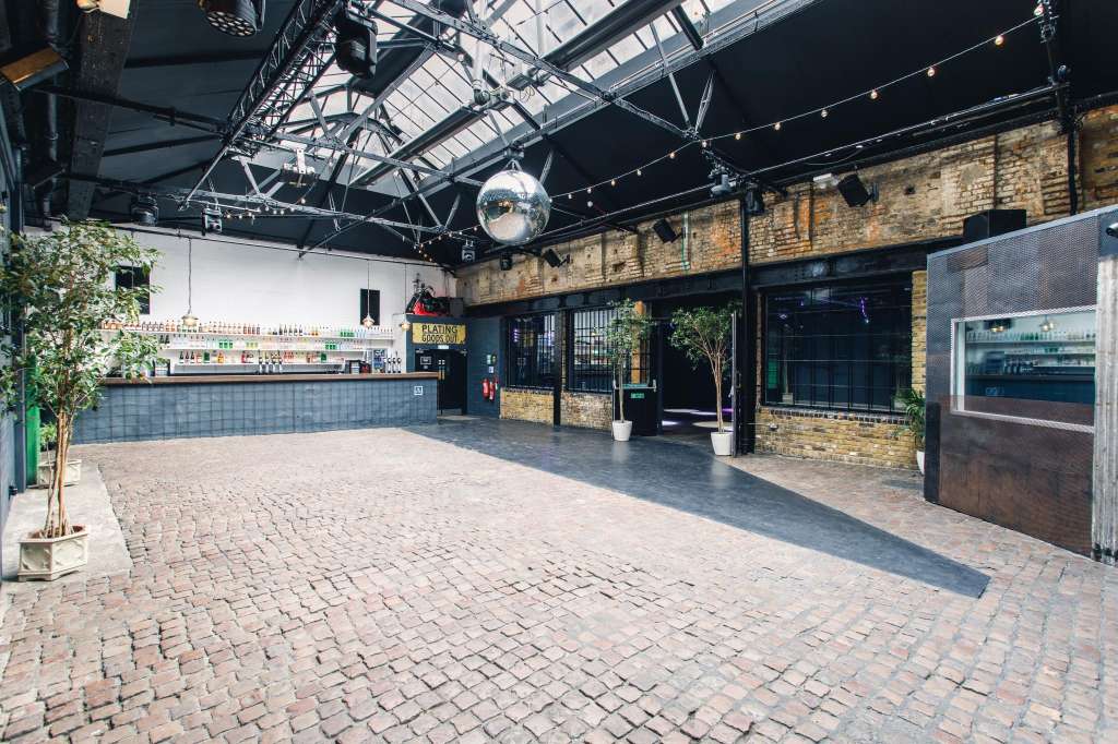 Industrial warehouse space with exposed brick, steel beams, bar counter, and disco ball under glass roof.