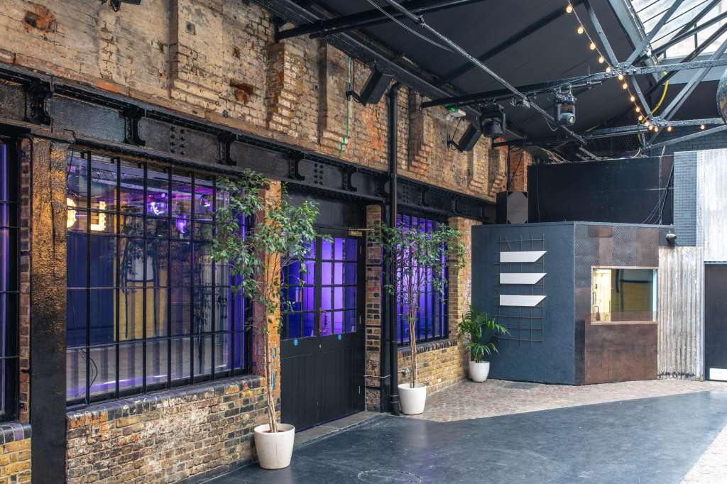Industrial venue exterior with exposed brick, purple-lit windows, potted plants, and steel structural elements.