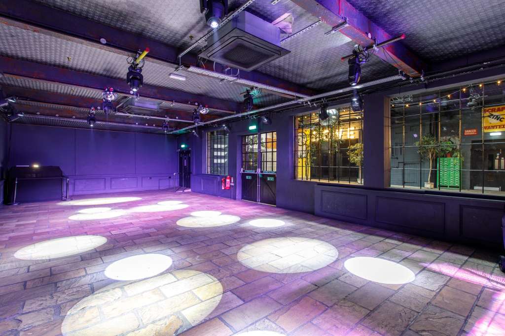 Industrial warehouse space with purple and pink ambient lighting, illuminated floor panels, and exposed ceiling infrastructure.