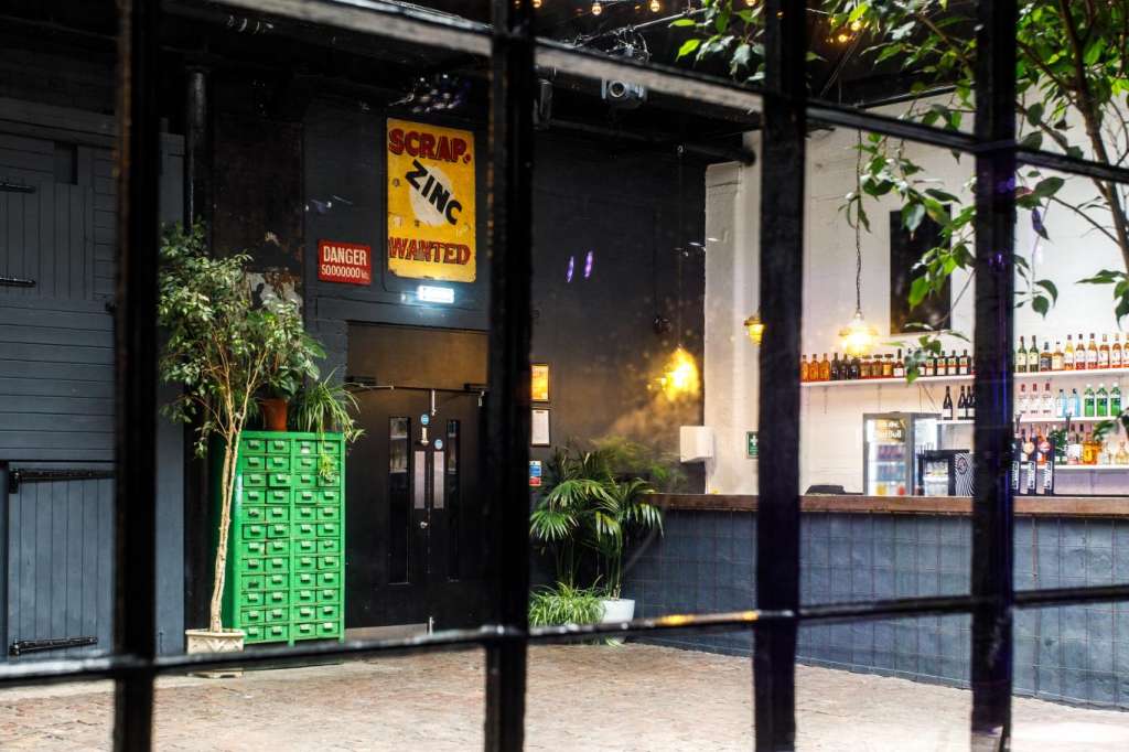 Industrial venue exterior with large disco ball, green door, bar counter, and plants visible through black-framed windows.
