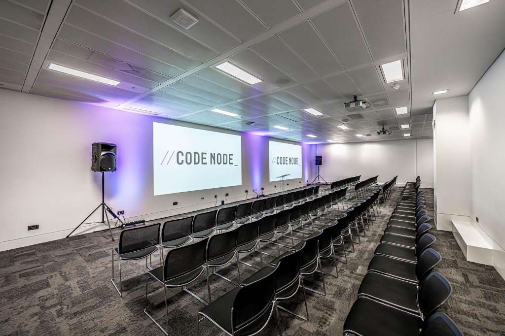 Conference room with rows of black chairs facing projection screens displaying CodeNode branding and purple ambient lighting.