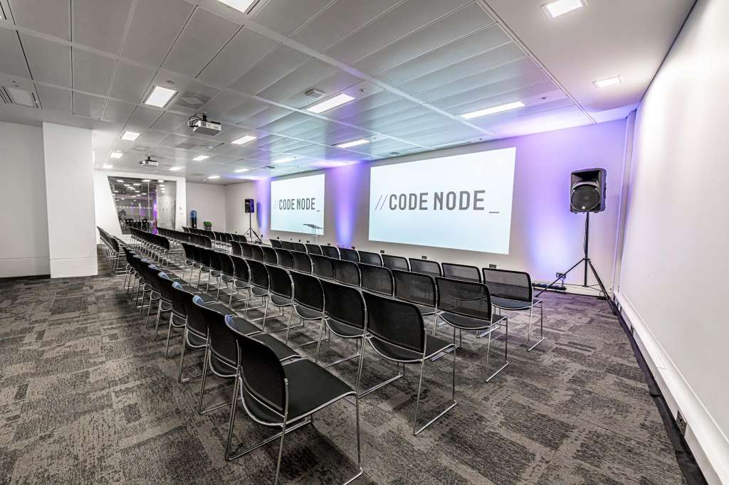 Conference room with rows of black chairs facing projection screens displaying CODE NODE branding and purple ambient lighting.