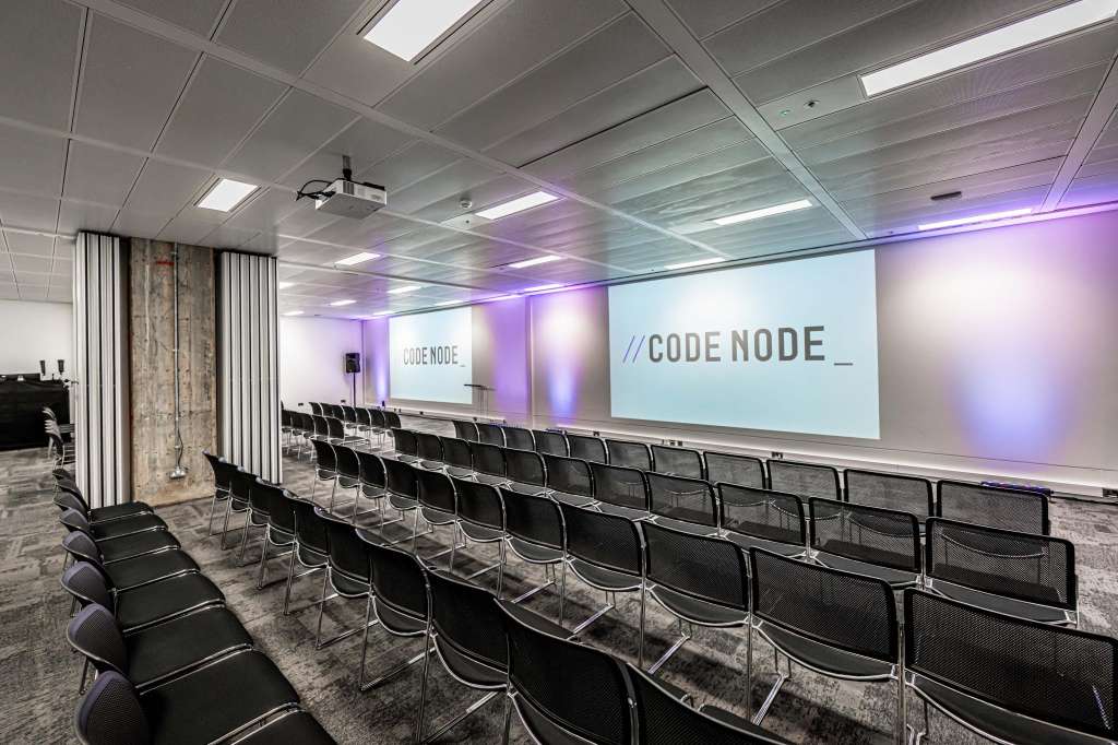 Auditorium with rows of black chairs facing a projection screen displaying CODE NODE branding, purple ambient lighting, and ceiling-mounted projector.