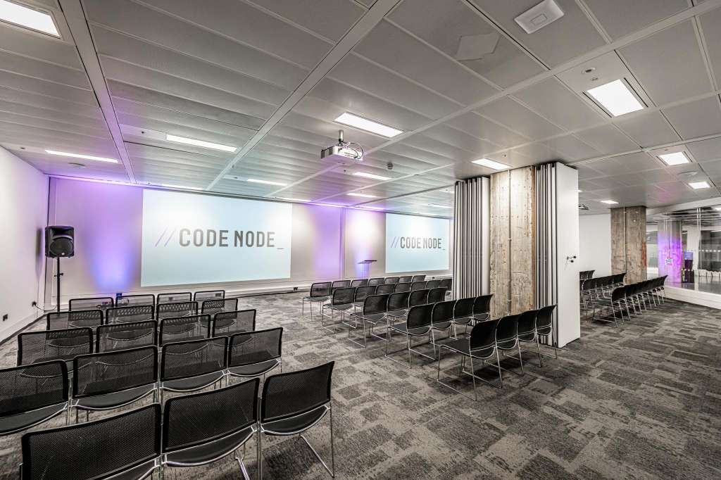 Modern event space with rows of black chairs facing projection screens displaying CODE NODE branding, purple accent lighting, and exposed wooden columns.