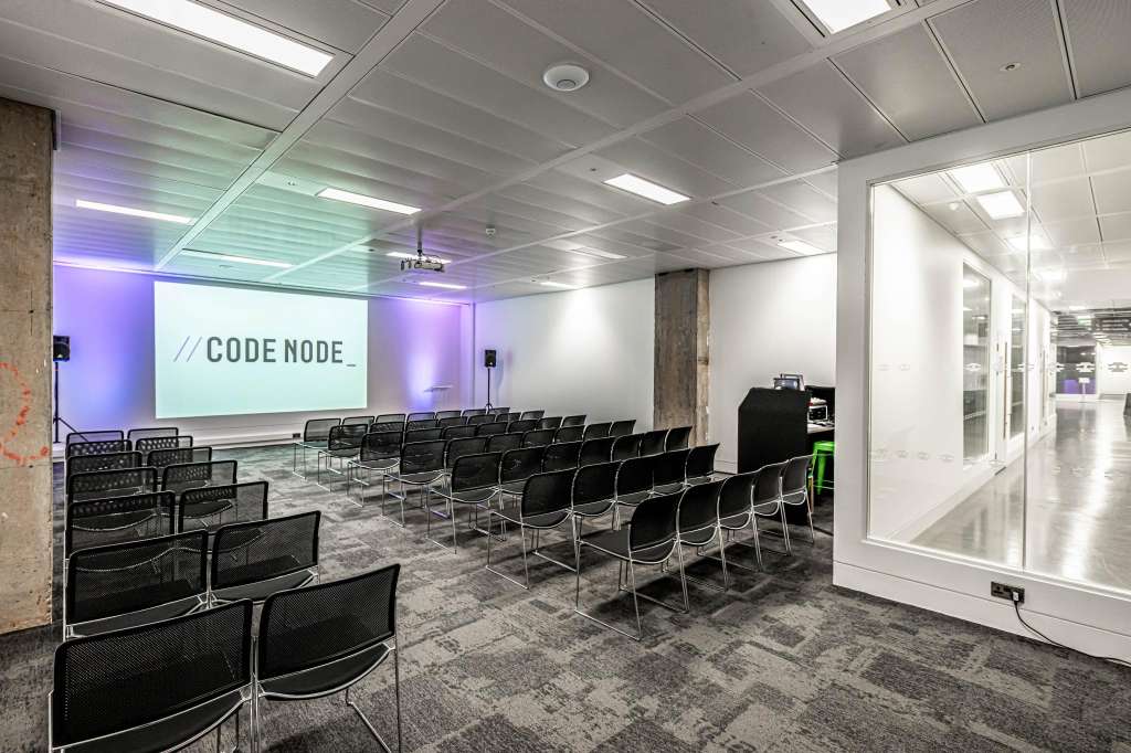 Modern conference room with rows of black chairs facing a projection screen displaying CodeNode branding, ambient purple lighting, and exposed brick wall.