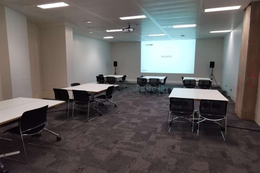 Modern meeting room with multiple white tables, black chairs, projector screen displaying Sony logo, and recessed lighting.