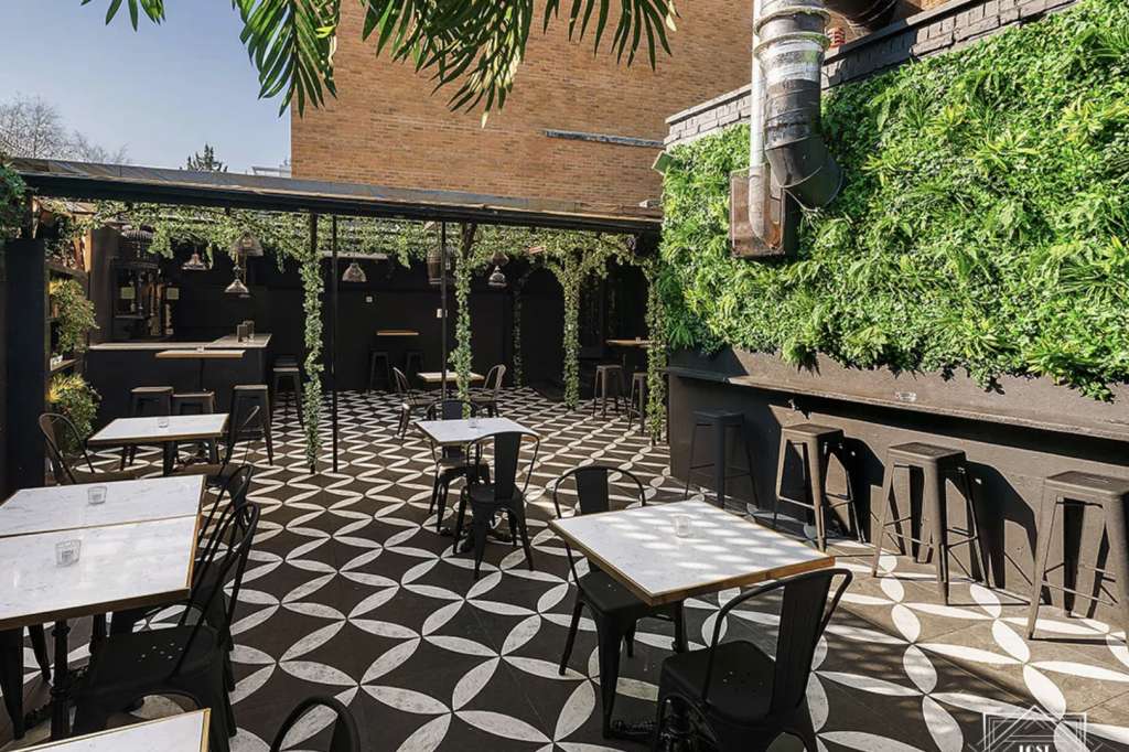 Outdoor terrace with geometric black and white flooring, scattered tables and chairs, ivy-covered walls, and green foliage overhead.
