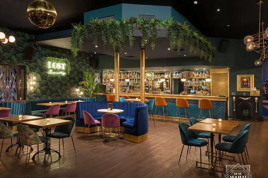 Main room with bar, colorful seating, hanging plants, and gold pendant lighting. Empty venue setup.