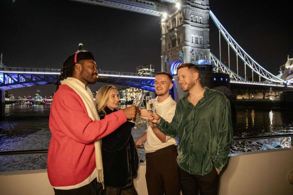 Four guests toasting with champagne glasses on an outdoor deck overlooking Tower Bridge at night.