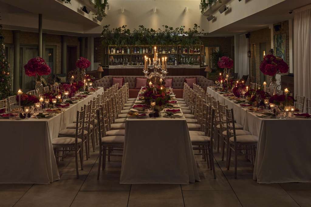 Dining room set for a formal seated dinner with long tables, burgundy floral centerpieces, candlelight, and festive garland.