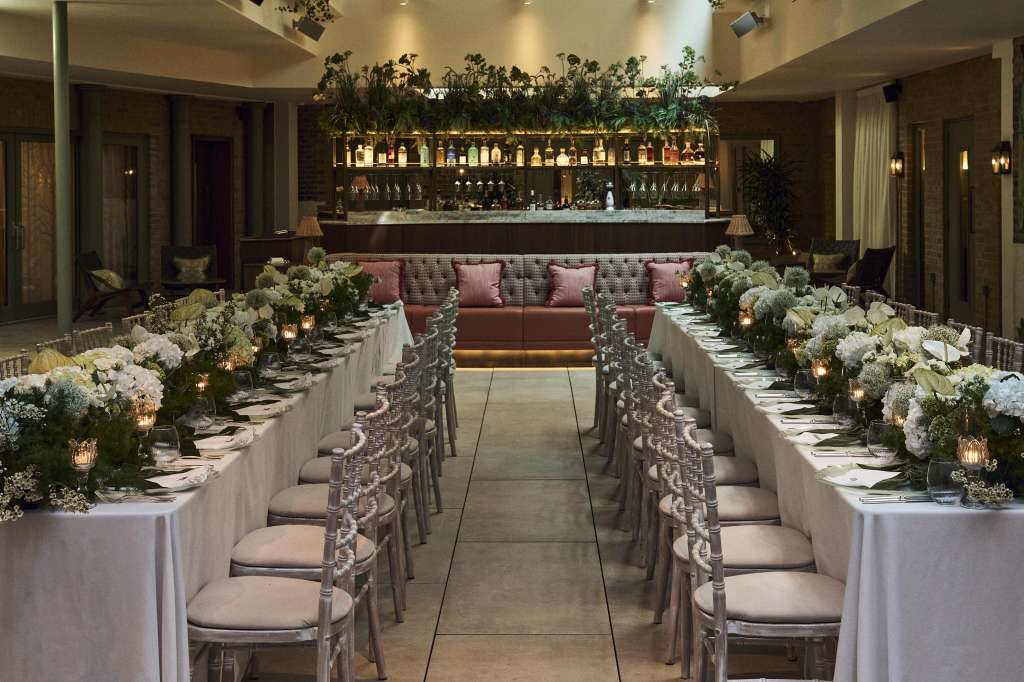 Dining room set for a formal seated dinner with long tables, white linens, floral centerpieces, and bar backdrop.
