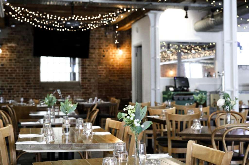 Warehouse dining space with long tables, wooden chairs, place settings, and string lights overhead.