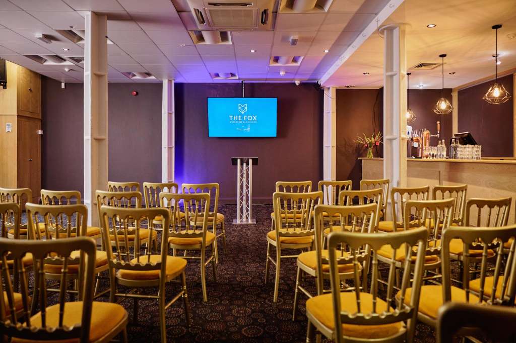 Event space with gold chairs arranged in rows facing a screen displaying THE FOX logo, purple ambient lighting, and bar counter.
