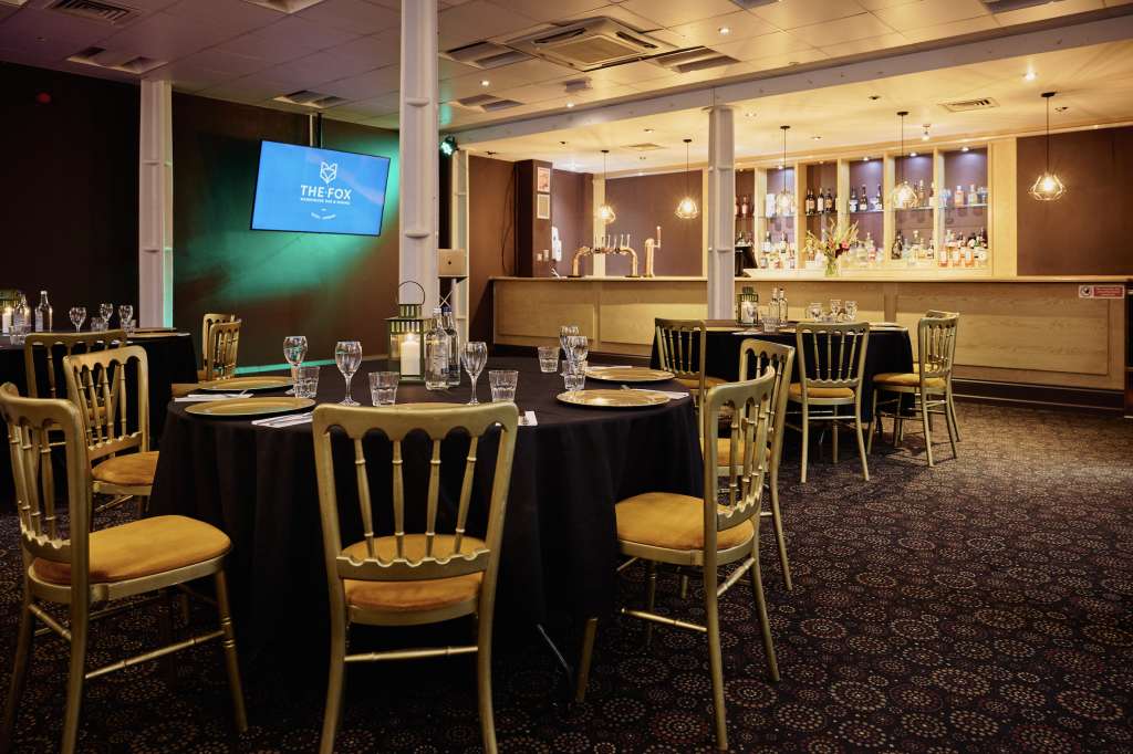 Dining room with round tables, black linens, gold chairs, and full bar with backlit shelving and green accent lighting.