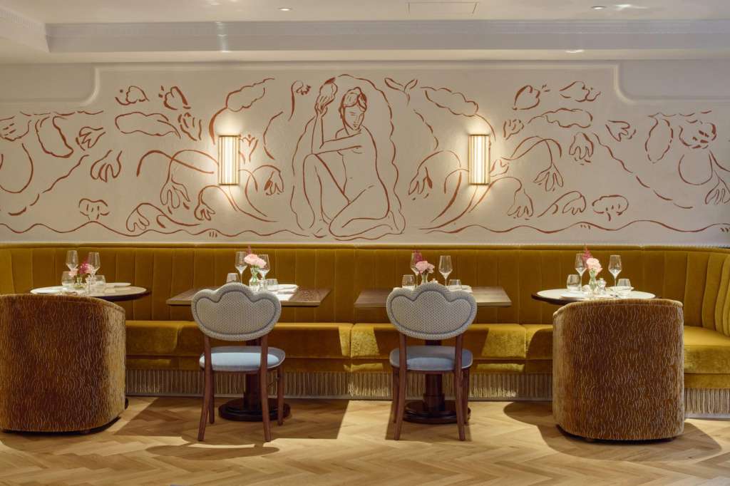 Dining room with banquette seating, round tables set with place settings, decorative wall mural, and warm lighting.