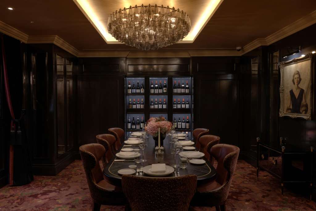 Private dining room with oval table, burgundy chairs, crystal chandelier, wine display, and portrait artwork.