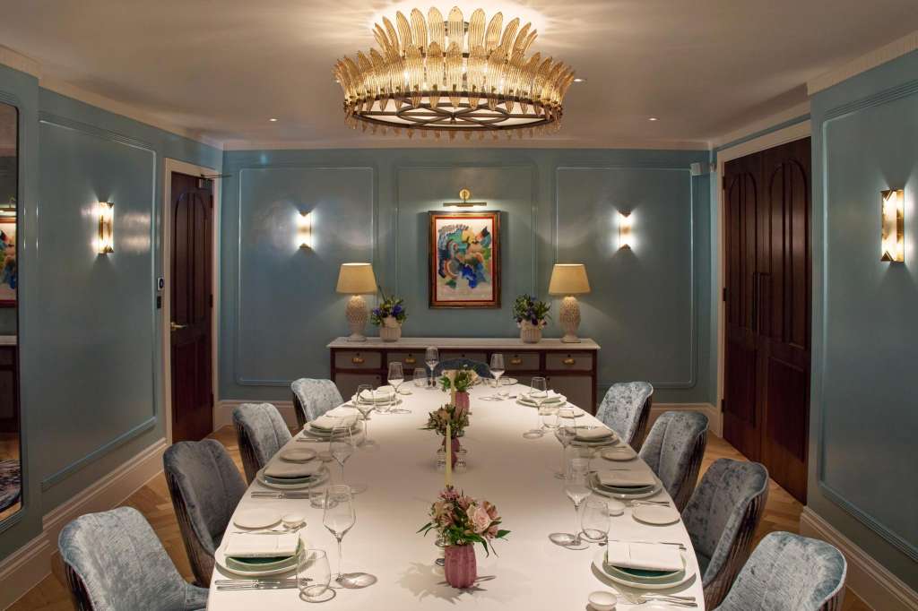 Elegant dining room with long white table, place settings, upholstered chairs, statement chandelier, and teal paneled walls.
