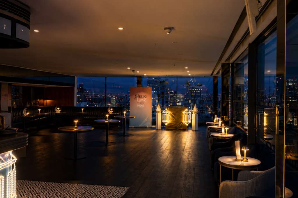 Rooftop bar interior at dusk with city skyline views, ambient lighting, high-top tables, and modern furnishings.