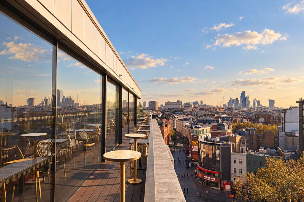Rooftop terrace with bar seating, floor-to-ceiling windows, and panoramic London cityscape at golden hour.